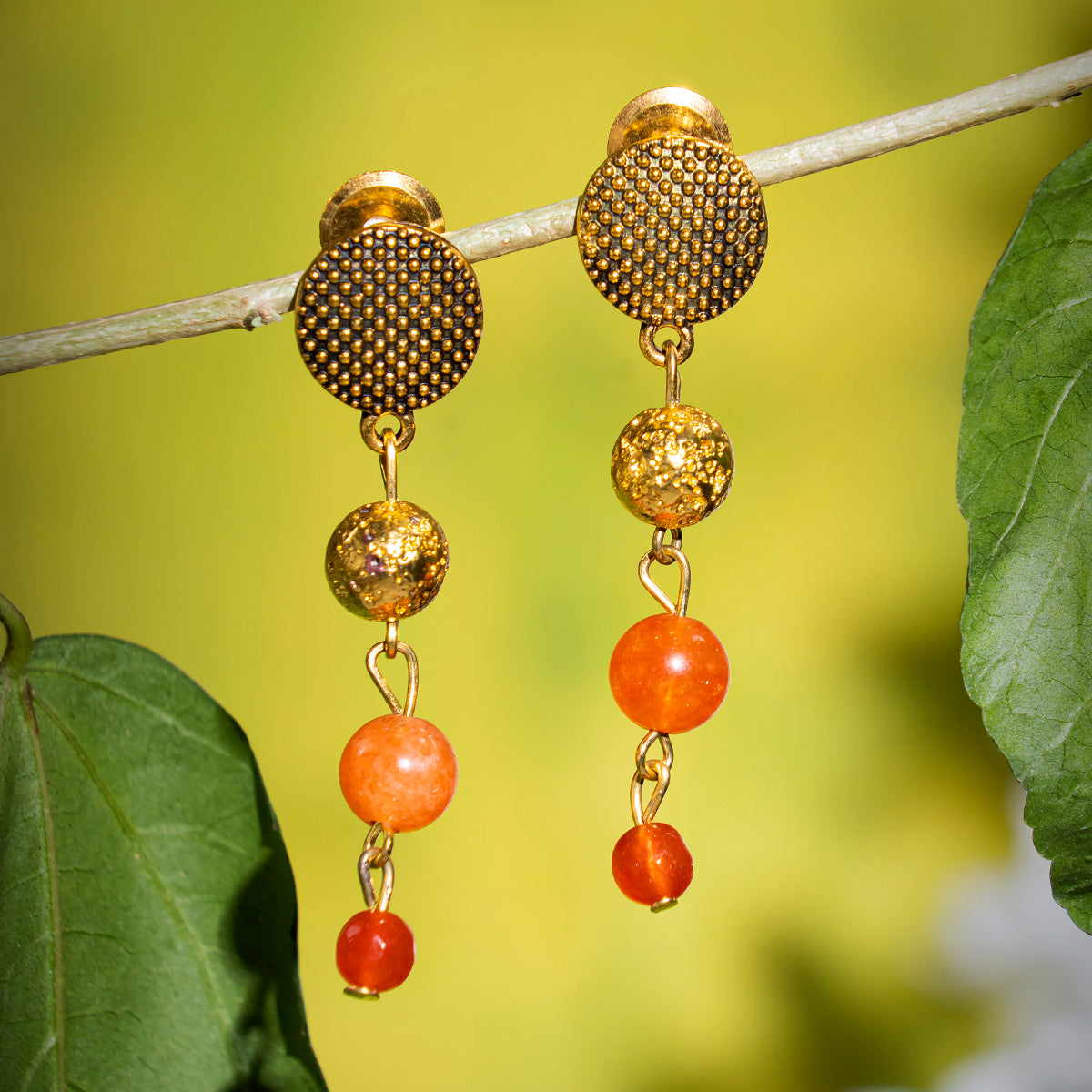 Eykaa earrings that has orange jade, golden lava semi precious stones hanging on a thin branch with green background