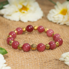 Eykaa EYKAA PINK JADE WITH SHELL PEARLS AND GOLDEN BEADS WITH GOLDEN CHARMS BRACELET on sack surface with white flowers.