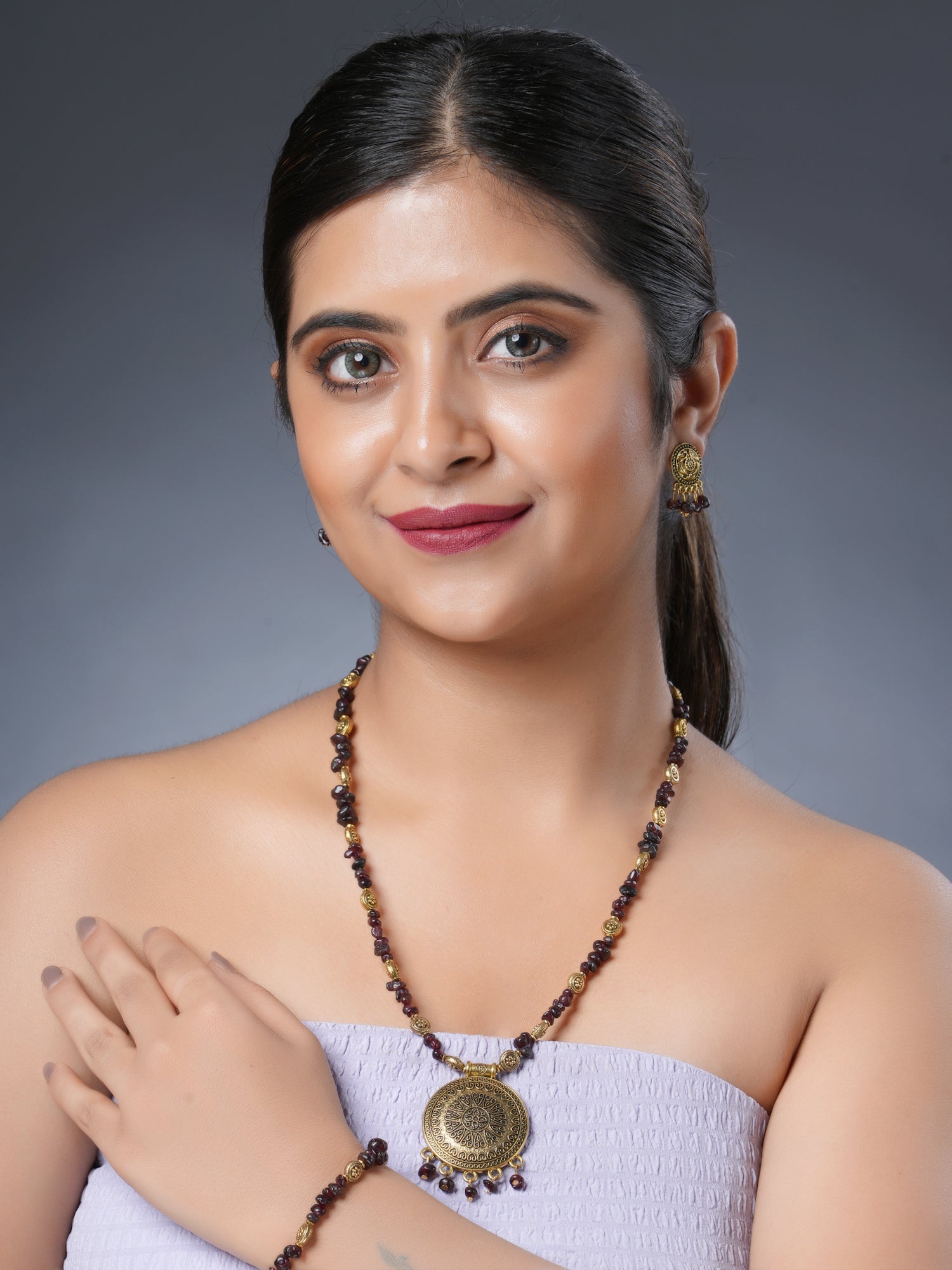 Girl model wearing Eykaa necklace, earrings and bracelet set that features garnet semi-precious stone with golden charms