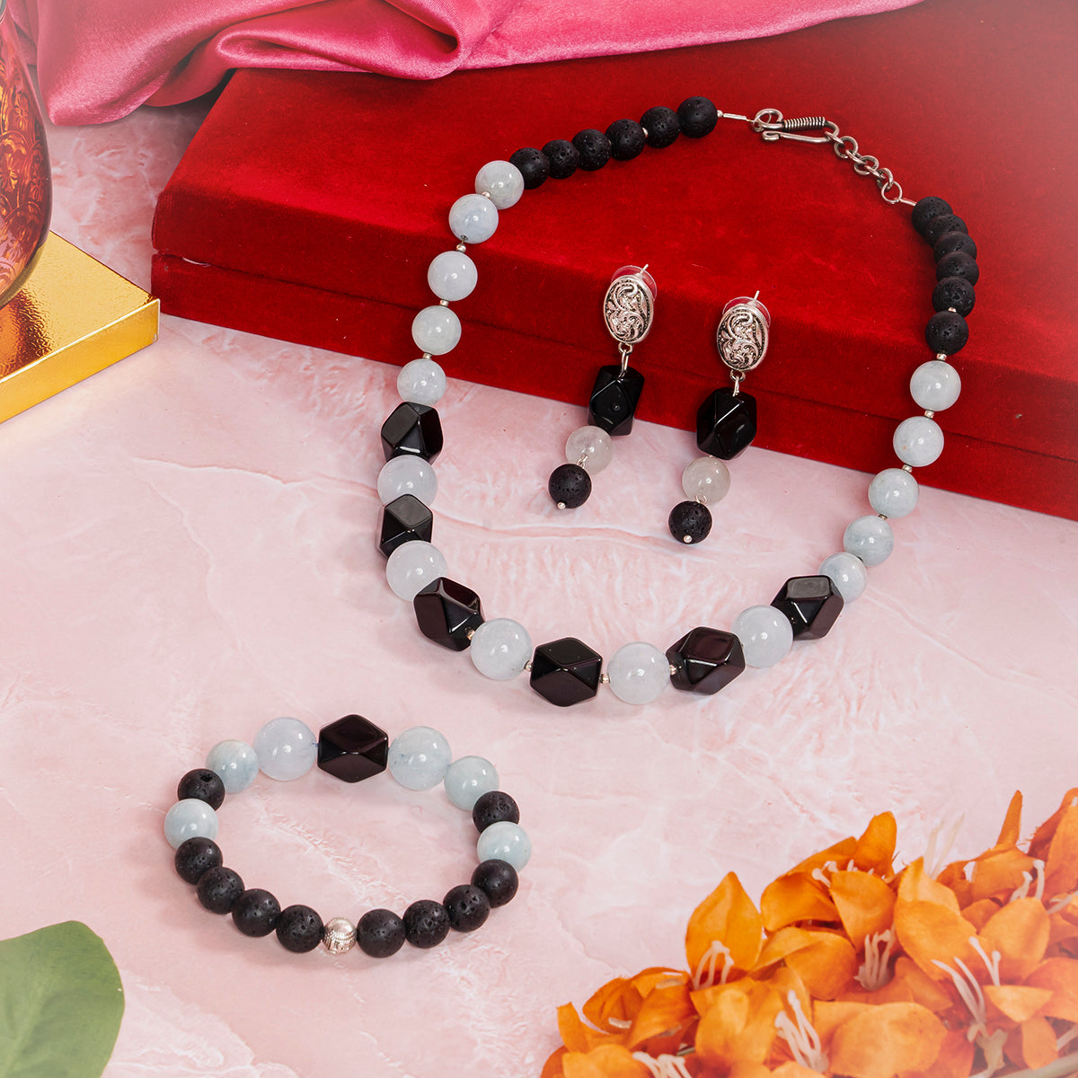 Eykaa Aquamarine With Lava And Black Onyx Necklace With Earrings And A Bracelet On A Soft Pink Marbled Surface, Arranged Around A Red Velvet Box, Surrounded By Orange Flowers And Green Leaves. 