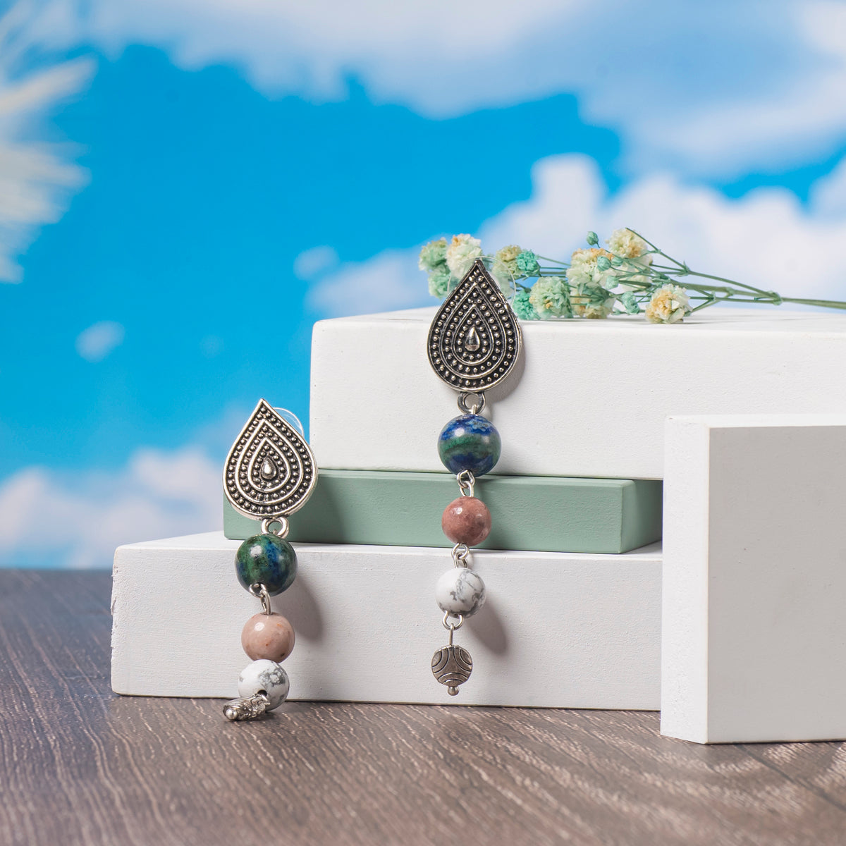  Eykaa Azurite, Pink Rhodonite And White Howlite Earrings Displayed On Multi-tiered Platform With Sky And Clouds Background.