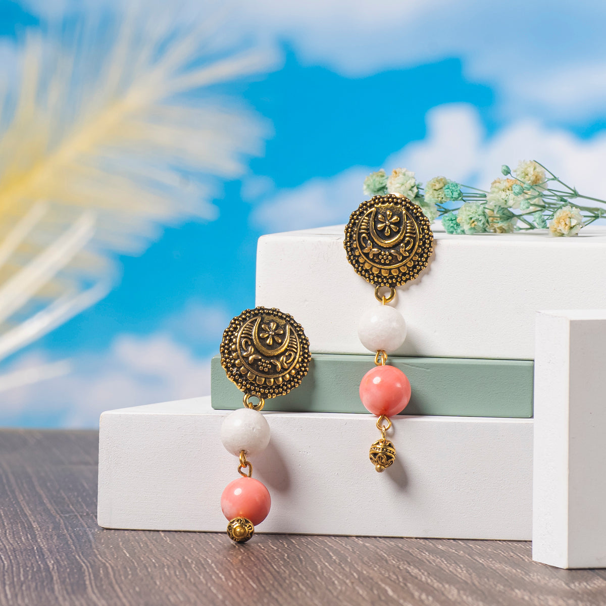 Eykaa Marble, Coral And Bamboo Coral With Golden Beads Earrings Displayed On Multi-tiered Platform With Sky And Clouds Background.