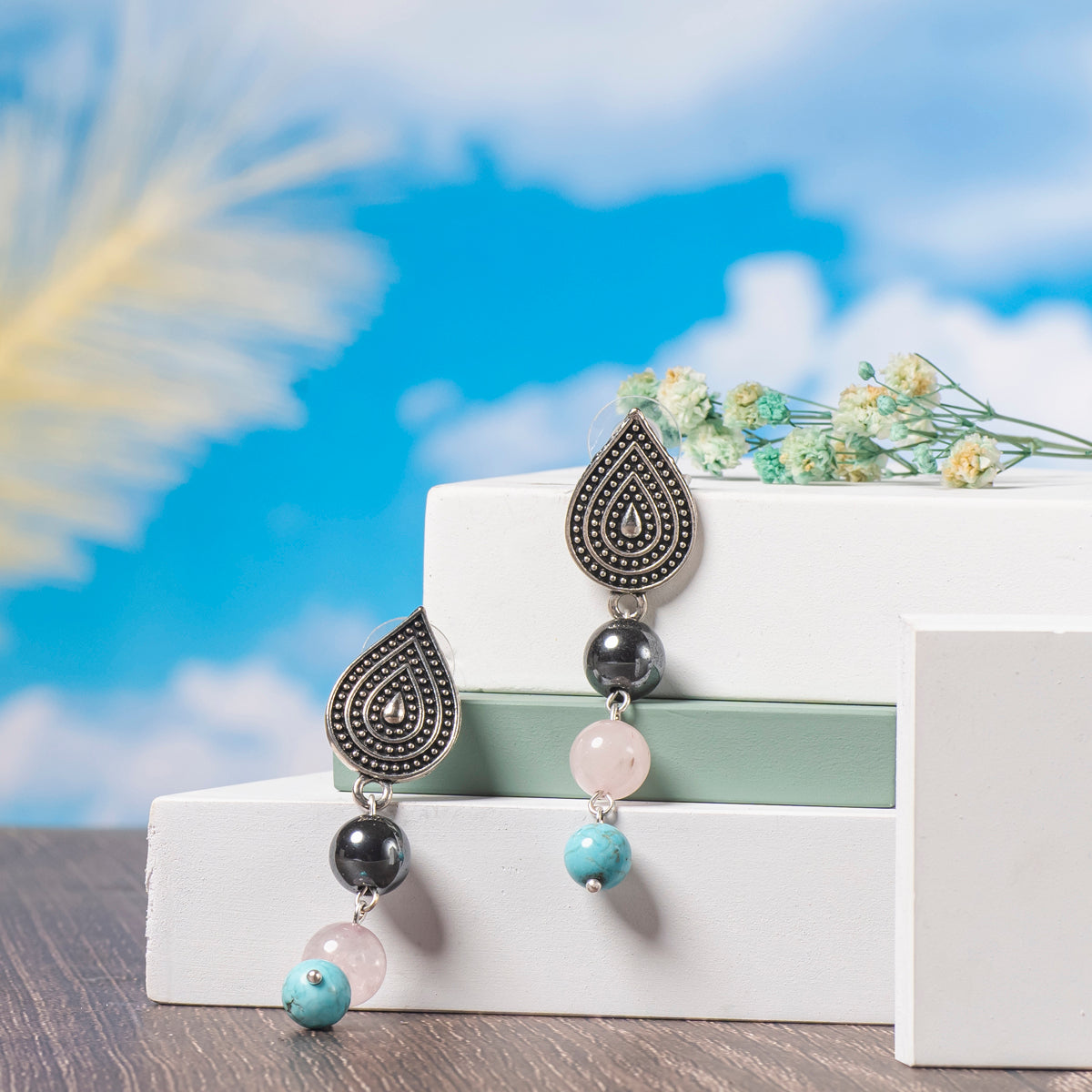 Eykaa Rose Quartz, Hematite And Marble With Firoza Earrings Displayed On Multi-tiered Platform With Sky And Clouds Background.