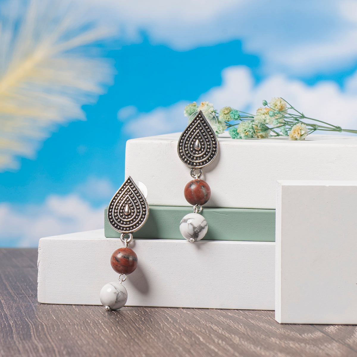 Eykaa Red Jasper, Silver Lava With White Howlite Earrings Displayed On Multi-tiered Platform With Sky And Clouds Background.