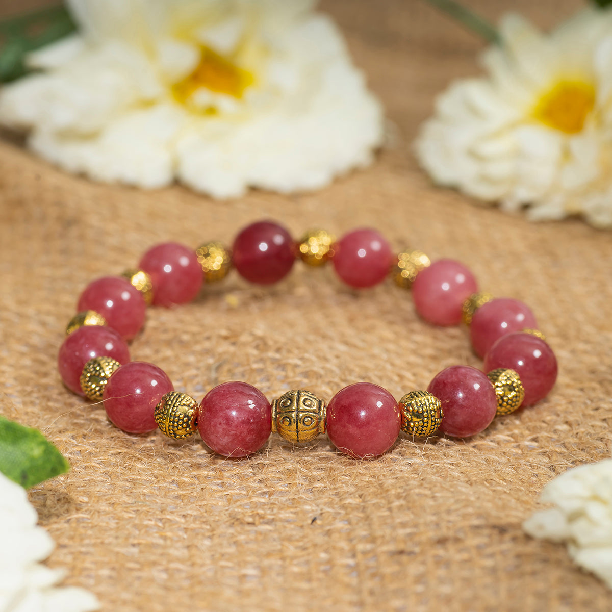 Eykaa EYKAA PINK JADE WITH SHELL PEARLS AND GOLDEN BEADS WITH GOLDEN CHARMS BRACELET on sack surface with white flowers.