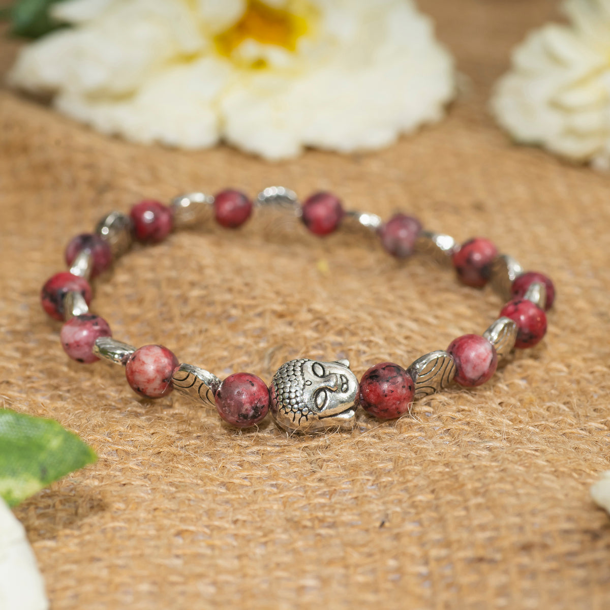 Eykaa EYKAA PINK DALMATIAN BRACELET WITH BUDDHA AND SILVER CHARMS on sack surface with white flowers.