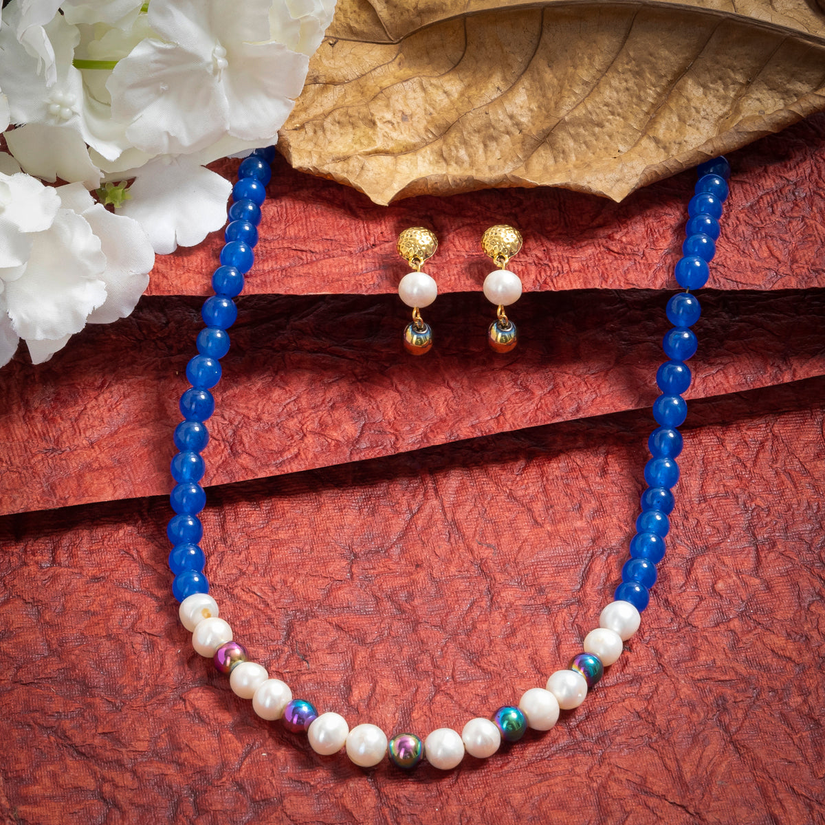 Eykaa Blue Onyx, Rainbow Hematite, And Freshwater Pearl Necklace With Earrings On A Textured Red Background With White Flowers.