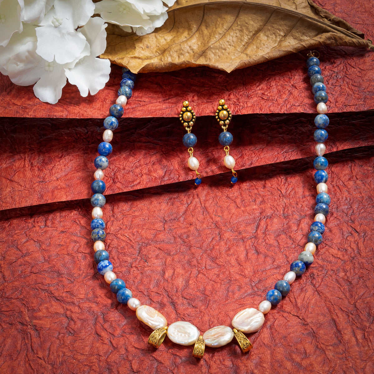 Eykaa Lapis Lazuli, Freshwater Pearl, And Jade Necklace With Earrings On A Textured Red Background With White Flowers.