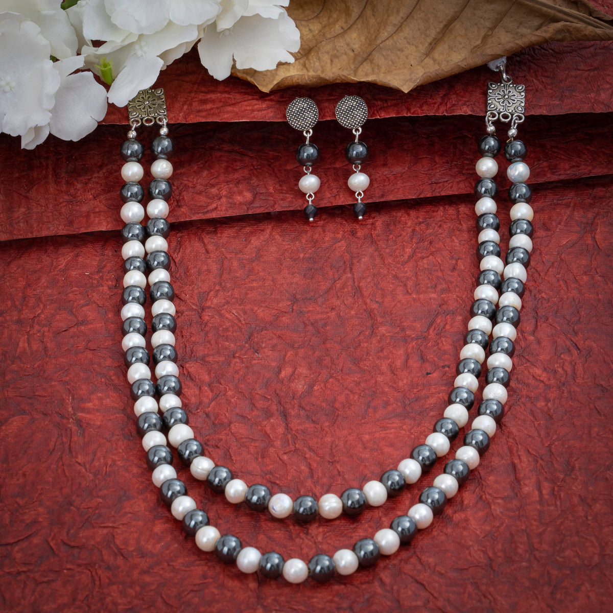 Eykaa Hematite And Freshwater Pearl Necklace With Earrings On A Textured Red Background With White Flowers.